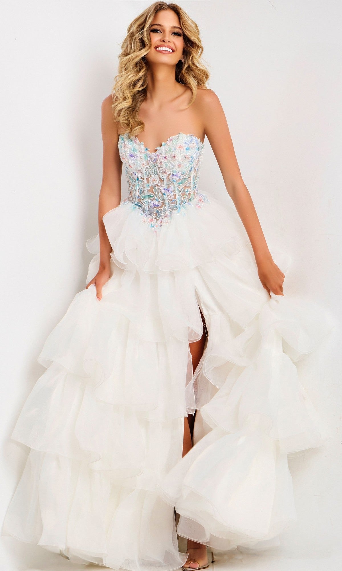 A woman smiles against a plain white background, holding the sides of her Jovani 48177 long prom dress— a strapless, ruffled white gown with a beaded corset bodice and pastel embellishments.